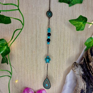 Könnte beinhalten: Ein kupferfarbenes Hängeornament mit türkisfarbenen und schwarzen Perlen, einem Schildkrötenanhänger und einem tropfenförmigen Anhänger. Das Ornament hängt an einer Kette und hat oben ein kreisförmiges Design. Der Hintergrund ist eine helle Holzstruktur mit Grün.