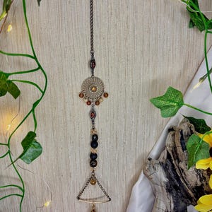 Puede incluir: Un adorno colgante decorativo con una cadena de bronce y detalles intrincados. El adorno presenta un diseño circular con cuentas de color ámbar, cuentas oscuras y un colgante triangular con un dije en forma de lágrima. El fondo incluye vegetación y flores.