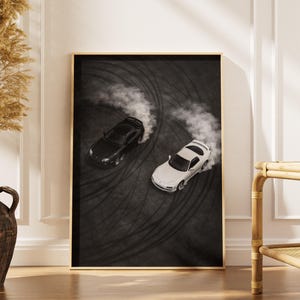 Könnte beinhalten: Gerahmter Kunstdruck mit zwei Sportwagen, die auf einer dunklen Rennstrecke driften und Rauch erzeugen. Ein Auto ist schwarz, das andere weiß. Das Bild ist in einem goldfarbenen Rahmen, geeignet für Wanddekoration.