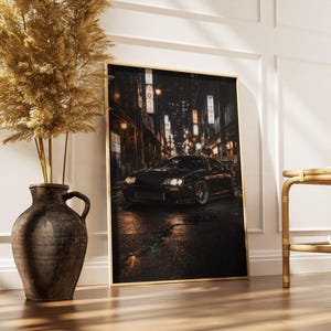 May include: A framed print featuring a black sports car parked on a wet street at night. The car is the focal point, with city lights and Japanese signage in the background. The print is displayed on a wooden floor, next to a decorative vase and a small table.
