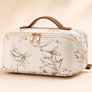 May include: A rectangular cosmetic bag in off-white featuring a dark brown botanical line art pattern. The bag includes a brown top handle, gold-toned metal zippers with dual pulls, and a structured design with rounded corners and piping.
