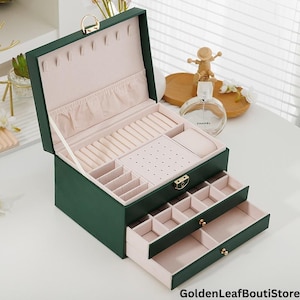 Può includere: Un portagioie verde scuro con più scomparti e cassetti. L'interno è foderato con un tessuto rosa chiaro. La scatola ha una chiusura e cerniere color oro. La parte superiore aperta rivela fessure per anelli e aree di stoccaggio.