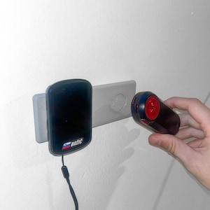 Puede incluir: Un dispositivo electrónico negro con la palabra "Bozic" impresa está montado en una pared. Un segundo dispositivo rojo y negro se sostiene cerca. Ambos dispositivos parecen ser parte de un sistema de seguridad o control de acceso.