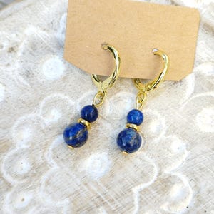 Peut inclure: Une paire de boucles d'oreilles créoles dorées avec des perles pendantes en lapis-lazuli bleu. Chaque boucle d'oreille comporte une petite perle dorée au-dessus d'une perle bleue ronde plus grande. Les boucles sont présentées sur une surface neutre.
