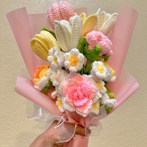 May include: A handmade crochet bouquet featuring a variety of colorful flowers. The bouquet includes white, yellow, pink, and orange flowers, wrapped in pink paper and tied with a white ribbon. The flowers are detailed and textured.