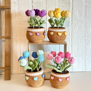 May include: Four crochet tulip arrangements in brown pots. Each pot has a white band with heart-shaped and round embellishments. The tulips are in shades of purple, yellow, blue, and pink, with green leaves. The pots sit on a shelf.