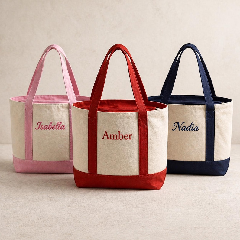 K&ouml;nnte beinhalten: Drei Canvas-Tragetaschen mit kontrastierenden Verzierungen und Griffen. Die Taschen sind in Rosa, Rot und Marineblau gehalten und tragen die Namen "Isabella", "Amber" und "Nadia" auf der Vorderseite. Die Taschen haben eine rechteckige Form.