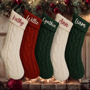 May include: Five cable-knit Christmas stockings in cream, red, and green hang in front of a fireplace. Each stocking is personalized with a name in a contrasting color: Kathy, Willie, Cynthia, Ann, and Liam.