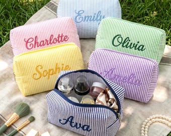 Könnte beinhalten: Eine Sammlung personalisierter Kosmetiktaschen in verschiedenen Pastellfarben, darunter Rosa, Gelb, Blau, Grün und Lila. Jede Tasche hat einen Namen, der in einer komplementären Farbe gestickt ist. Eine Tasche ist geöffnet und zeigt Kosmetikprodukte.
