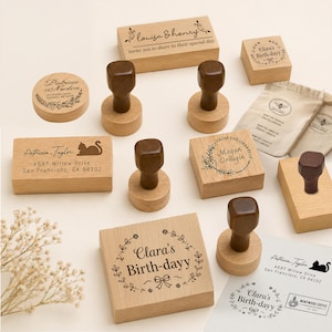 May include: A collection of wooden stamps with various designs and text. Includes stamps with names, addresses, and phrases like "Clara's Birth-dayy" and "Louisa & Henry". The stamps have brown handles and are arranged on a light surface.