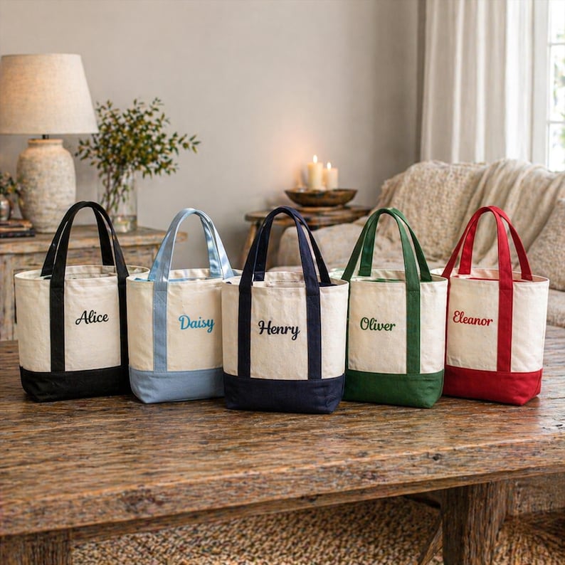 K&ouml;nnte beinhalten: F&uuml;nf Canvas-Tragetaschen mit farblich abgesetzten B&ouml;den und Griffen. Jede Tasche hat einen anderen Namen auf der Vorderseite gestickt: Alice, Daisy, Henry, Oliver und Eleanor. Die Taschen stehen auf einem Holztisch.