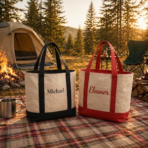 K&ouml;nnte beinhalten: Zwei Canvas-Tragetaschen mit schwarzen und roten Akzenten, personalisiert mit Namen. Eine Tasche hat schwarze Griffe und den Namen "Michael" in Schwarz. Die andere hat rote Griffe und den Namen "Eleanor" in Rot. Die Taschen liegen auf einer karierten Decke in der N&auml;he eines Campingplatzes.