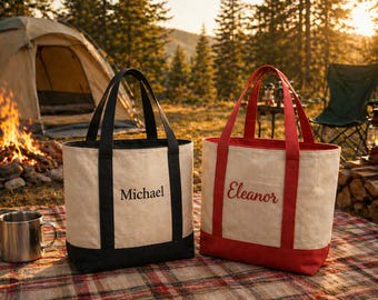 Könnte beinhalten: Zwei Canvas-Tragetaschen mit schwarzen und roten Akzenten, personalisiert mit Namen. Eine Tasche hat schwarze Griffe und den Namen "Michael" in Schwarz. Die andere hat rote Griffe und den Namen "Eleanor" in Rot. Die Taschen liegen auf einer karierten Decke in der Nähe eines Campingplatzes.