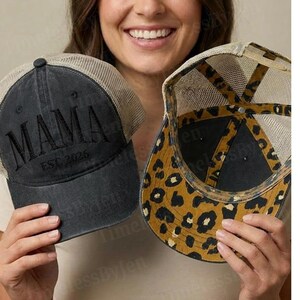 May include: Two trucker hats. One is dark gray with "MAMA EST 2026" embroidered on the front, a tan mesh back, and a black brim. The other hat has a leopard print pattern on the underside of the brim and a black interior.