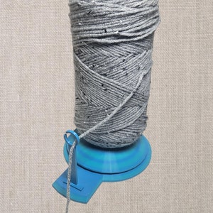 May include: A blue yarn caddy with a gray ball of yarn on top. The yarn caddy has a swivel base and a yarn guide.