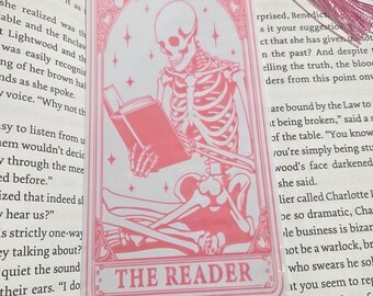 Può includere: Segnalibro rosa con uno scheletro che legge un libro, con la scritta "THE READER". Il segnalibro ha un nappa rosa ed è posto sopra un libro aperto.