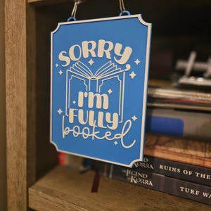 May include: A blue sign with white text that reads "SORRY I'M FULLY BOOKED" hangs from a shelf. The sign has a decorative border and features an open book graphic with stars. The sign is hanging in front of a bookshelf.