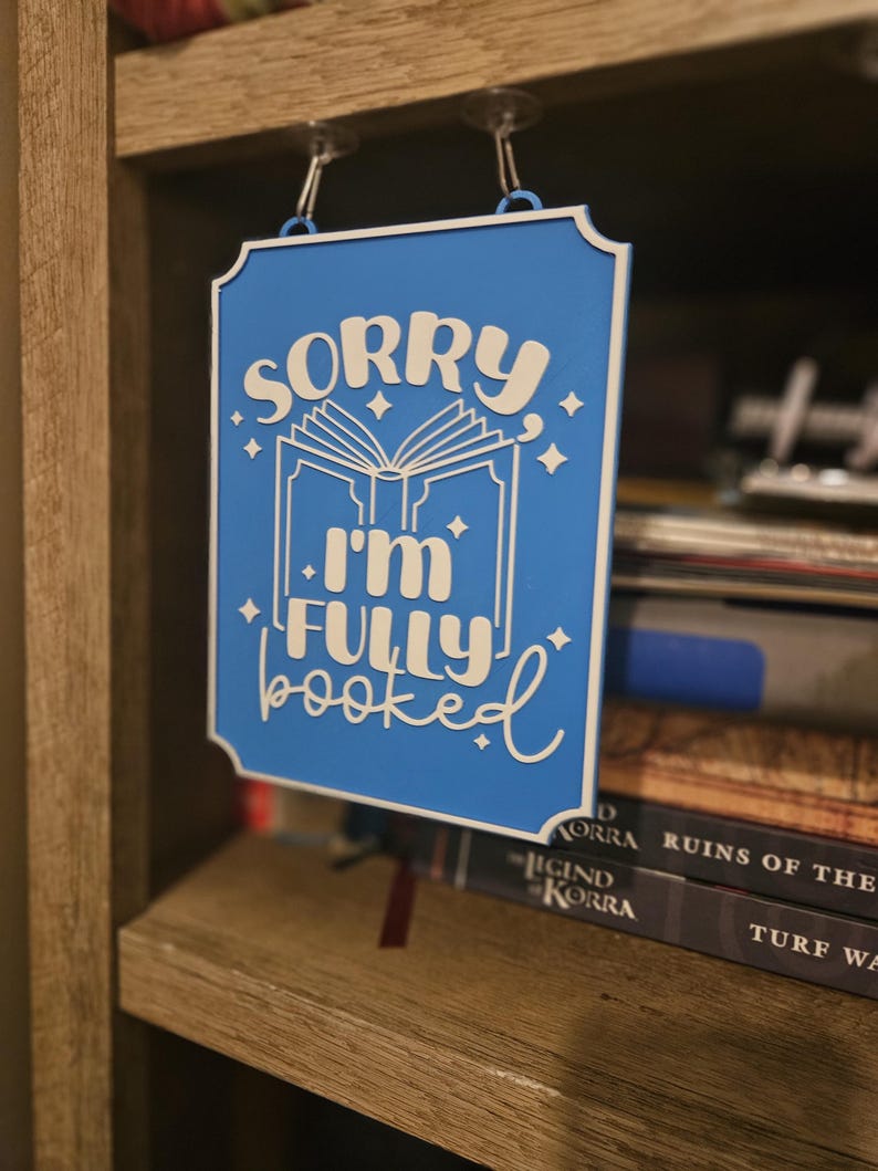 May include: A blue sign with white text that reads "SORRY I'M FULLY BOOKED" hangs from a shelf. The sign has a decorative border and features an open book graphic with stars. The sign is hanging in front of a bookshelf.