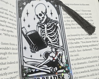 Può includere: Segnalibro in bianco e nero con uno scheletro che legge un libro, con la scritta "THE READER". Il segnalibro ha un nappa nera e un bordo decorativo con stelle. Il tema generale è gotico e mistico.