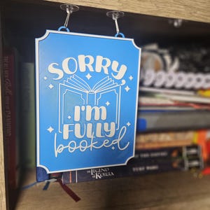 May include: A blue rectangular sign with the text "SORRY I'M FULLY booked" in white script. The sign is hanging from a shelf with clear suction cups. The sign is placed in front of a bookshelf filled with books.