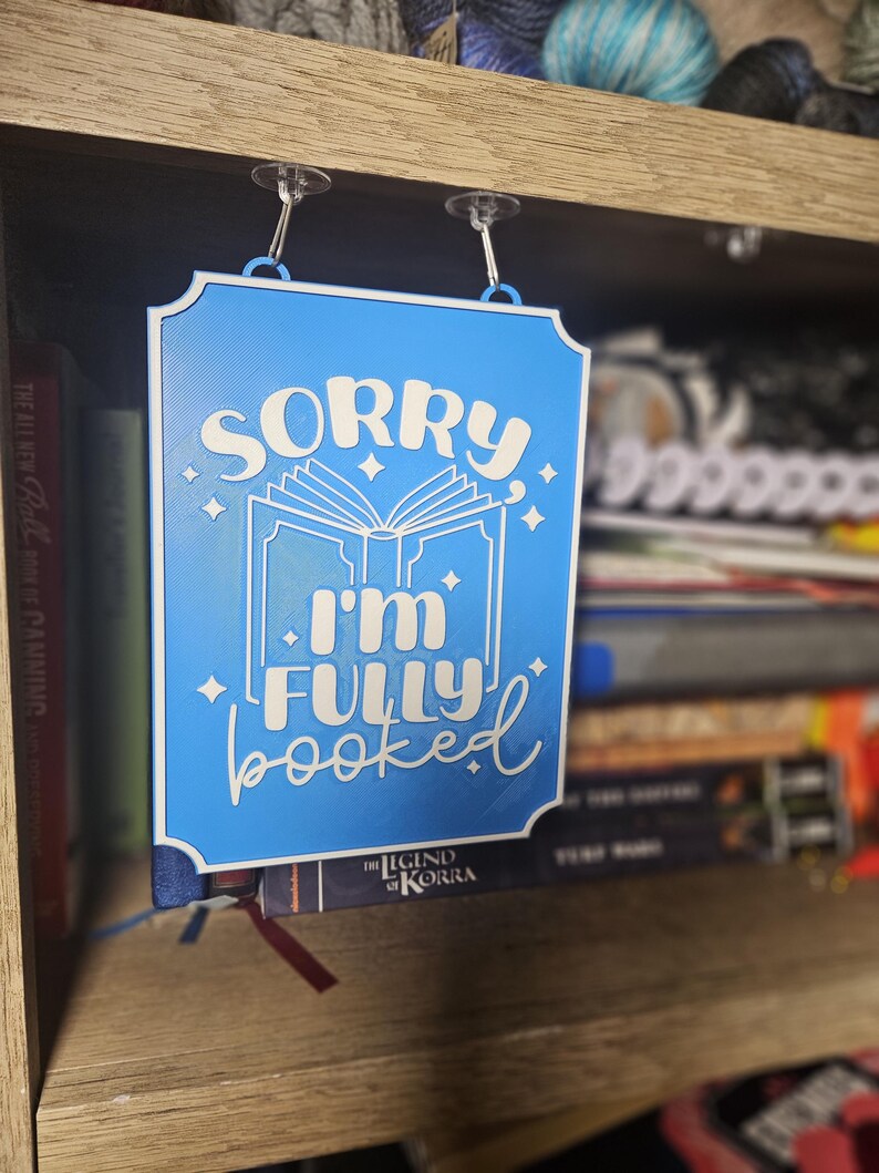 May include: A blue rectangular sign with the text "SORRY I'M FULLY booked" in white script. The sign is hanging from a shelf with clear suction cups. The sign is placed in front of a bookshelf filled with books.
