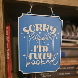 May include: A blue wooden sign with white lettering that reads "SORRY I'M FULLY booked". The sign features an open book graphic and star accents. It is hanging on a shelf with books in the background.