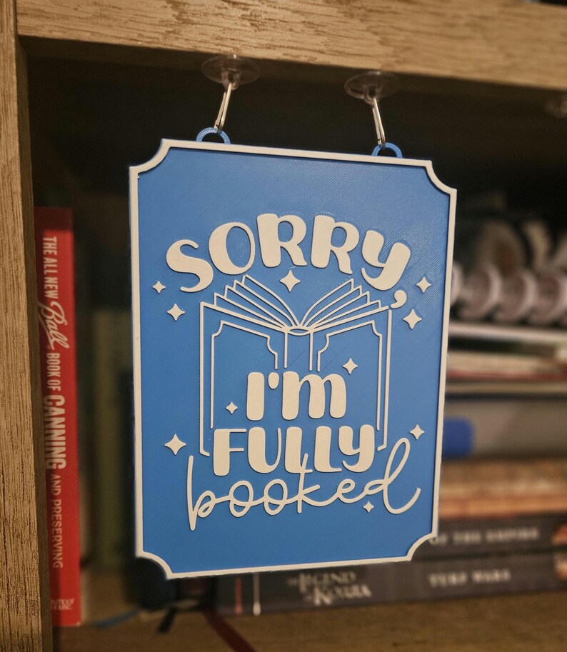 May include: A blue wooden sign with white lettering that reads "SORRY I'M FULLY booked". The sign features an open book graphic and star accents. It is hanging on a shelf with books in the background.
