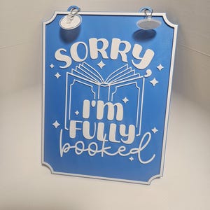 May include: Blue rectangular sign with white text that reads "SORRY I'M FULLY booked". The sign has a white border and a graphic of an open book. The sign has two suction cups attached to the top.