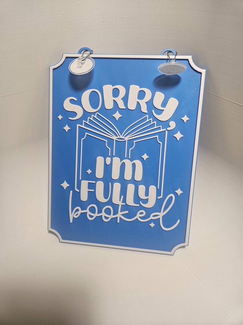 May include: Blue rectangular sign with white text that reads "SORRY I'M FULLY booked". The sign has a white border and a graphic of an open book. The sign has two suction cups attached to the top.