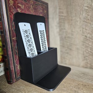 Puede incluir: Un soporte para marcapáginas de madera negra con dos marcapáginas blancos cortados con láser. Los marcapáginas tienen intrincados diseños geométricos.