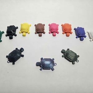 May include: A collection of small, colorful turtle figurines in various colors, including green, black, yellow, brown, pink, orange, blue, and white. The turtles are arranged in two rows, with some featuring a small loop.