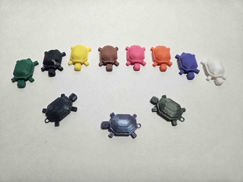 May include: A collection of small, colorful turtle figurines in various colors, including green, black, yellow, brown, pink, orange, blue, and white. The turtles are arranged in two rows, with some featuring a small loop.