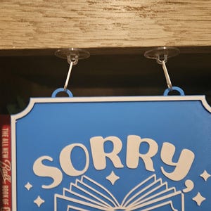 May include: A blue sign with white text that reads "SORRY" above an open book illustration. The sign has a white border and is suspended by two clear suction cups.