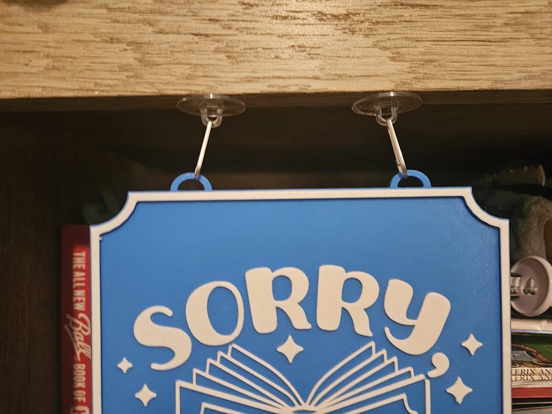 May include: A blue sign with white text that reads "SORRY" above an open book illustration. The sign has a white border and is suspended by two clear suction cups.