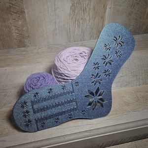 May include: A dark blue sock-shaped knitting tool with a snowflake design and rows of small holes. Two balls of yarn, one pink and one purple, are in the background.