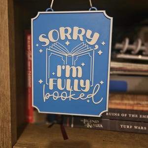 May include: A blue sign with white text that reads "SORRY I'M FULLY BOOKED". The sign has a white border and is hanging from two clear glass hooks. The sign is in front of a bookshelf.