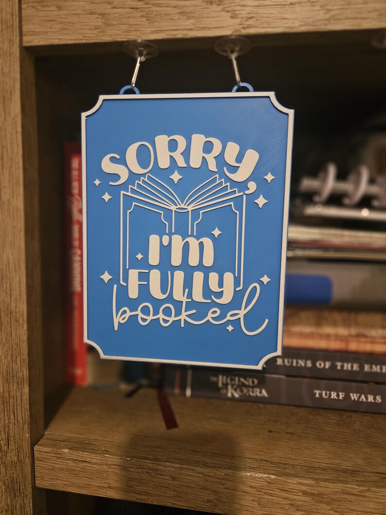 May include: A blue sign with white text that reads "SORRY I'M FULLY BOOKED". The sign has a white border and is hanging from two clear glass hooks. The sign is in front of a bookshelf.