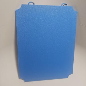 May include: A rectangular, light blue sign with rounded corners and two small, looped handles at the top. The sign has a textured surface and a solid blue color.