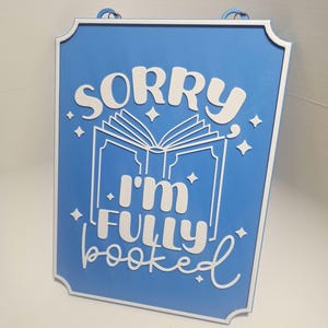 May include: A rectangular blue sign with white text and a white border. The sign reads "SORRY I'M FULLY booked" with an open book graphic. The sign has a rope for hanging.