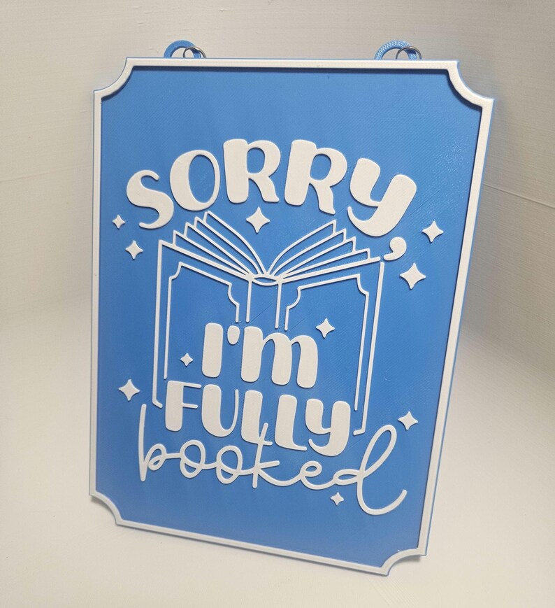 May include: A rectangular blue sign with white text and a white border. The sign reads "SORRY I'M FULLY booked" with an open book graphic. The sign has a rope for hanging.