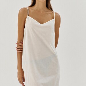 May include: A white cotton slip dress with thin straps. The dress has a V-neckline and a straight silhouette.