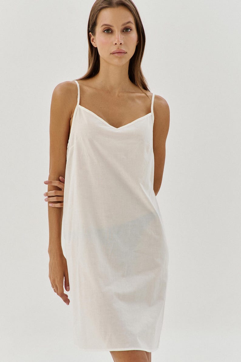 May include: A white cotton slip dress with thin straps. The dress has a V-neckline and a straight silhouette.