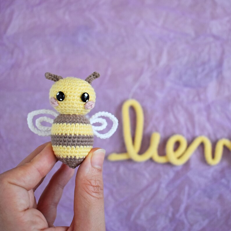 May include: A crocheted bee with yellow and brown stripes, white wings, and large black eyes. The bee has small antennae and pink cheeks. The word "bee" is written in yellow in the background. The bee is held in a person's hand.