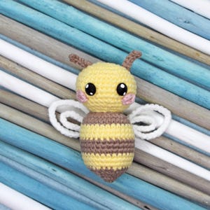 May include: A handmade crochet bee with a yellow head and body, brown stripes, and white wings. The bee has large black eyes and pink cheeks. The toy is set against a background of blue, white, and gray wooden sticks.