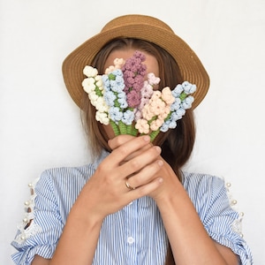 以下が含まれることがあります： 麦わら帽子をかぶった人が、顔の前にかぎ針編みで作った花束を持っています。花はピンク、青、白の色合いです。