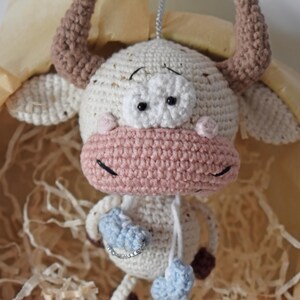 May include: A crocheted white cow with brown horns, pink nose, and blue and white accents. The cow is hanging from a string.