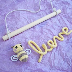 May include: A handmade crochet baby mobile featuring a yellow and brown bee, the word "love" in yellow, and a wooden dowel. The bee has white wings and is suspended by white string against a purple background.