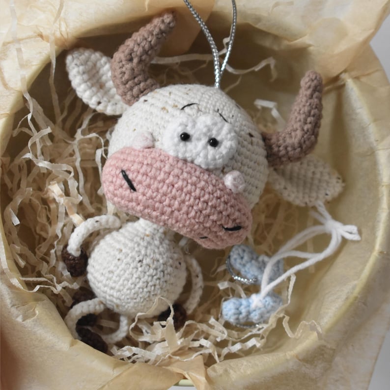 May include: A crocheted white cow with brown horns and pink nose, hanging from a string. The cow is sitting in a basket filled with shredded paper.