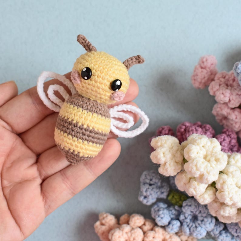 May include: A handmade crochet bee with yellow and brown stripes, white wings, and black eyes. The bee is held in a hand, with a light blue background. Crochet flowers in various colors are in the background.