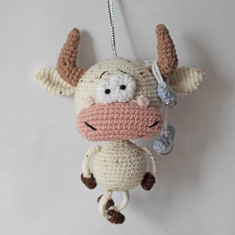 May include: A crocheted white cow with brown horns and pink nose, hanging from a silver string. The cow has black eyes and a white background.
